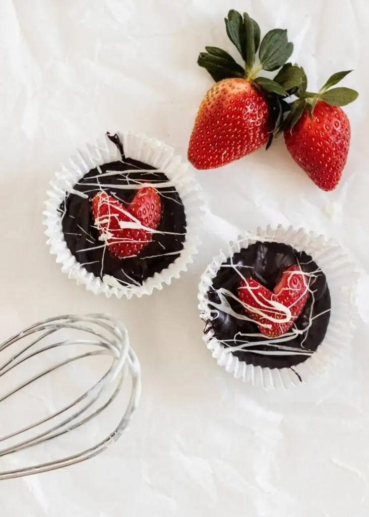 Two rich chocolate cups filled with glossy dark chocolate, topped with fresh strawberry slices and drizzled with white chocolate, presented in paper liners. This elegant Valentine's Day dessert is a delightful indulgence for fans of chocolate sweets and romantic treats.