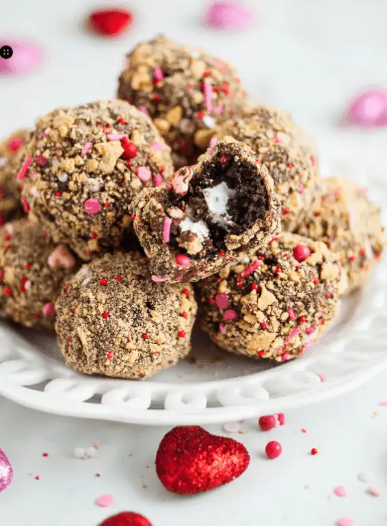 A plate of festive chocolate truffles coated in crushed graham crackers, sprinkles, and red and pink accents, with one truffle bitten to reveal a creamy marshmallow center. This playful Valentine's Day dessert is a delightful indulgence for chocolate sweets lovers, perfect for celebrating the season of love.