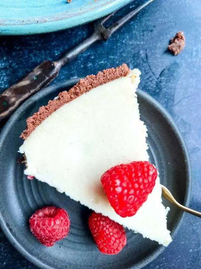 A slice of creamy cheesecake with a chocolate crust, garnished with fresh raspberries, served on a dark plate with scattered berries nearby. This decadent Valentine's Day dessert combines the richness of chocolate sweets with a light, fruity touch for a romantic treat.