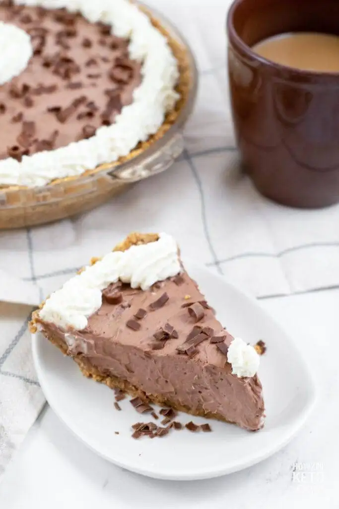 A slice of silky chocolate cream pie with a graham cracker crust, topped with whipped cream and sprinkled with chocolate shavings, served on a white plate. The whole pie and a cup of coffee sit in the background, making this Valentine's Day dessert a delightful choice for chocolate sweets lovers.