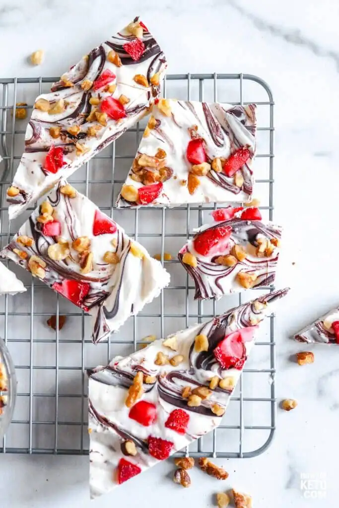 Pieces of creamy white chocolate bark swirled with dark chocolate, topped with vibrant red strawberries and crunchy walnuts, arranged on a cooling rack. This festive Valentine's Day dessert is a visually stunning and irresistible treat for chocolate sweets lovers.