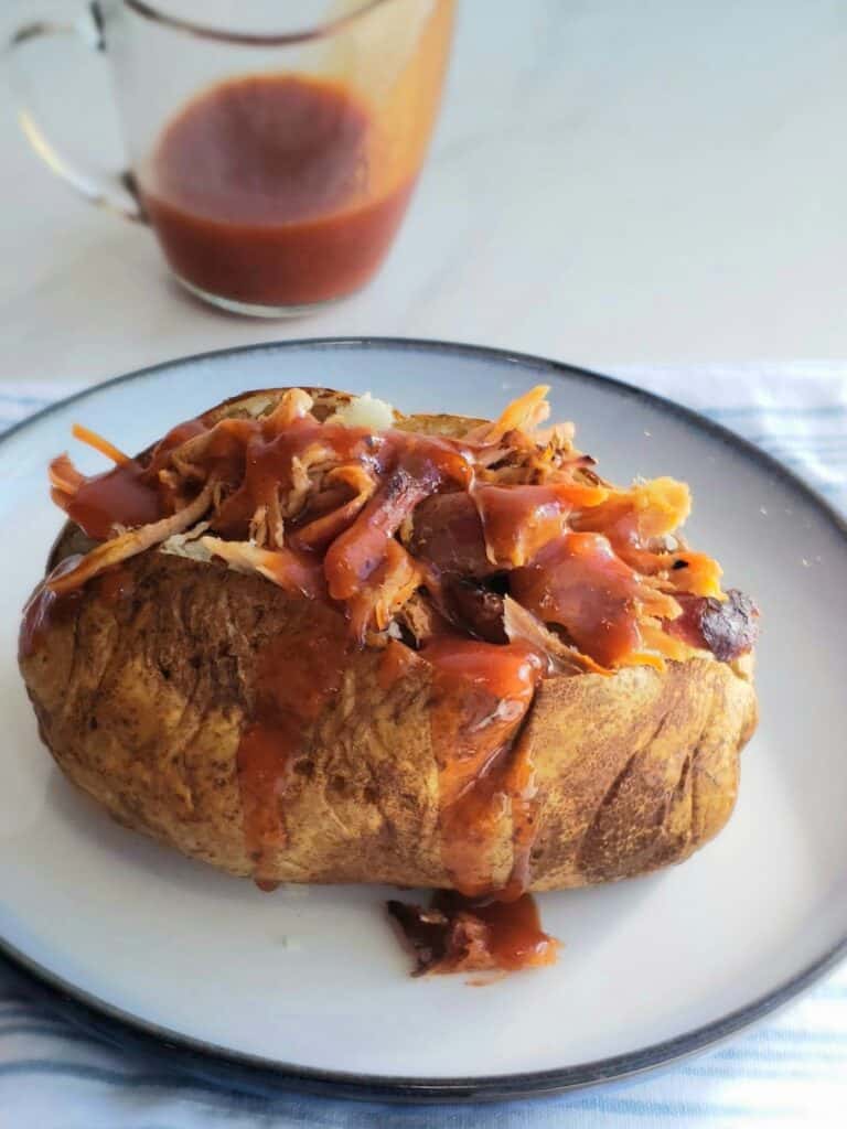 Baked potato loaded with leftover pulled pork and drizzled with barbecue sauce, served on a white plate with a pitcher of sauce in the background.