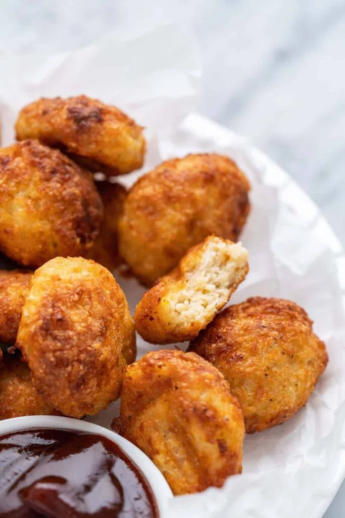 Golden, crispy chicken nuggets arranged on parchment paper, with a small bowl of barbecue sauce in the corner. A kid-friendly and crowd-pleasing Super Bowl food idea for game day.