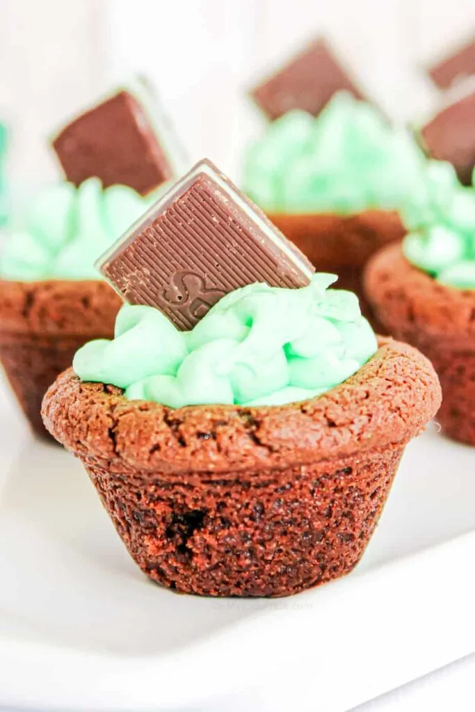 Chocolate brownie cups filled with creamy green mint frosting and topped with a chocolate square, perfect for St Patrick's Day desserts or festive Irish treats.