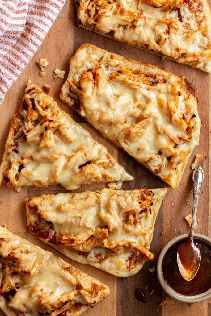 Slices of BBQ chicken flatbread topped with shredded chicken, melted cheese, and a rich BBQ sauce, served on a wooden board. A small bowl of extra BBQ sauce and a striped napkin accompany the dish, making it a tasty and shareable Superbowl appetizer.
