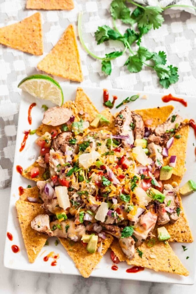 A plate of Hawaiian BBQ chicken nachos arranged with crispy tortilla chips, tender BBQ chicken, diced pineapple, chopped red onion, avocado, shredded cheese, and a drizzle of BBQ sauce. Garnished with fresh cilantro and served with a lime wedge, this tropical twist on nachos is a flavorful Superbowl appetizer.