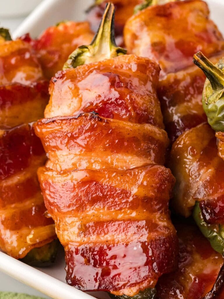 Glazed bacon-wrapped jalapeño poppers stuffed with a creamy filling, baked to perfection and served on a white platter. These smoky, spicy, and savory bites are a crowd-pleasing Superbowl appetizer.