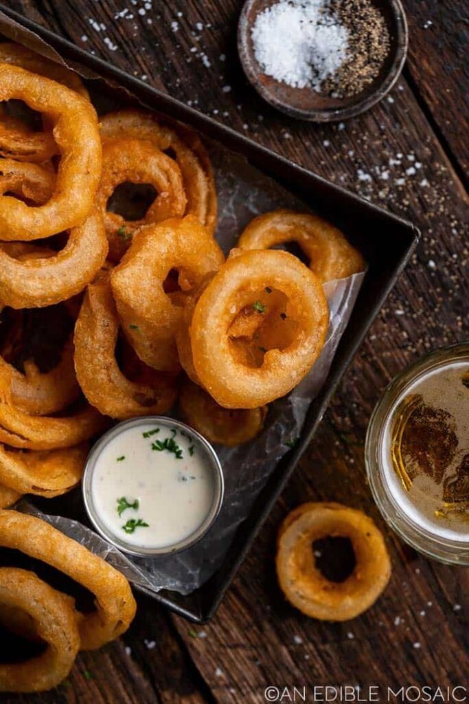 A basket of crispy beer-battered onion rings served with a creamy dipping sauce, accompanied by a small bowl of salt and pepper for seasoning. Set on a rustic wooden table with a glass of beer, these indulgent treats are a classic Superbowl appetizer.