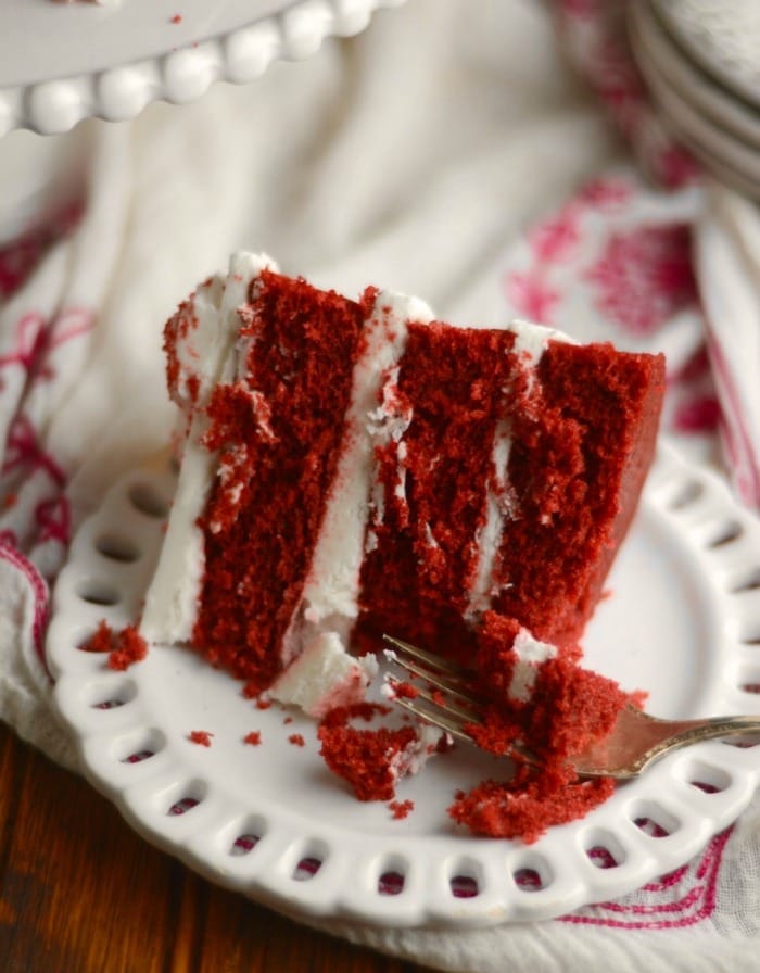 Close-up of a slice of vibrant red velvet cake with creamy white frosting, served on a decorative white plate, making it a timeless and romantic choice for Valentine's Cake Ideas.