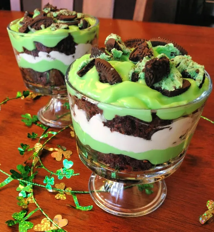 Layered St Patrick's Day trifle in glass bowls featuring green pudding, chocolate cake, whipped cream, and crushed mint Oreos, decorated with shamrock confetti for a festive Irish dessert.