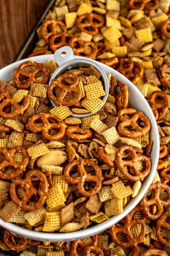 A white bowl filled with a mix of pretzels, Chex cereal, and nuts, with a scoop resting on top, surrounded by more snack mix on a tray. A crunchy and savory Super Bowl food idea perfect for sharing.