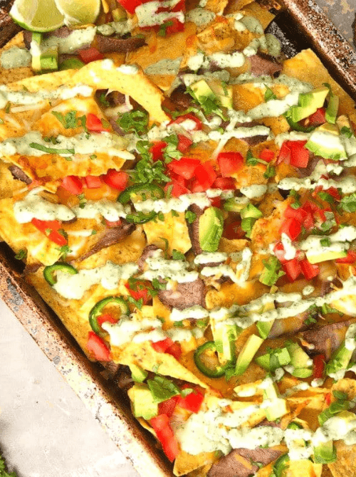 Loaded sheet pan nachos topped with leftover steak, melted cheese, diced tomatoes, avocado, jalapeños, and drizzled with creamy sauce, garnished with fresh cilantro and lime wedges.