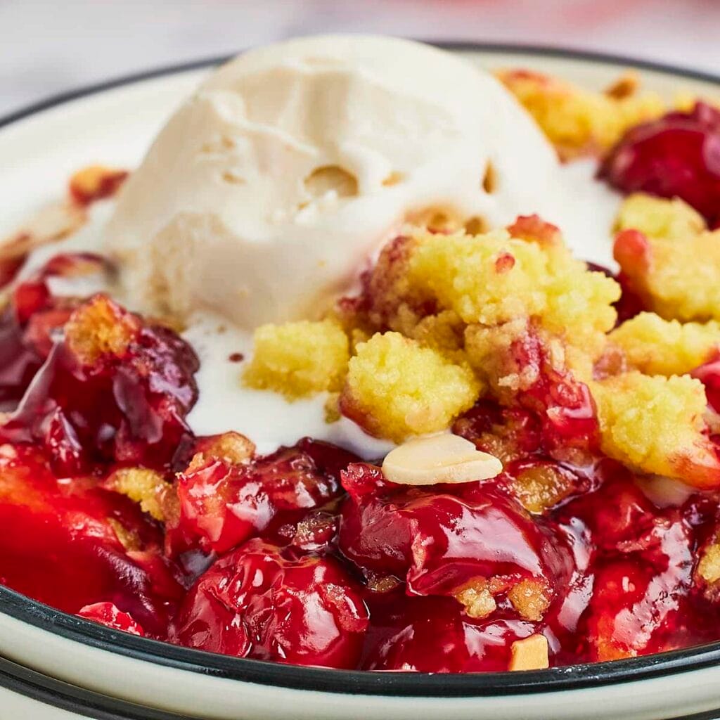 Close-up of a warm cherry crumble dessert topped with a scoop of vanilla ice cream, offering a unique twist for Valentine's Cake Ideas with its vibrant red cherries and golden crumble topping.