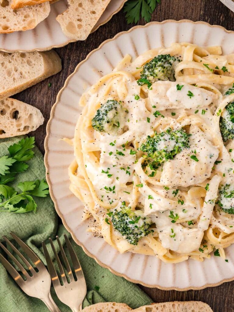 A plate of creamy pasta topped with leftover chicken and broccoli, garnished with fresh herbs and served alongside slices of crusty bread.