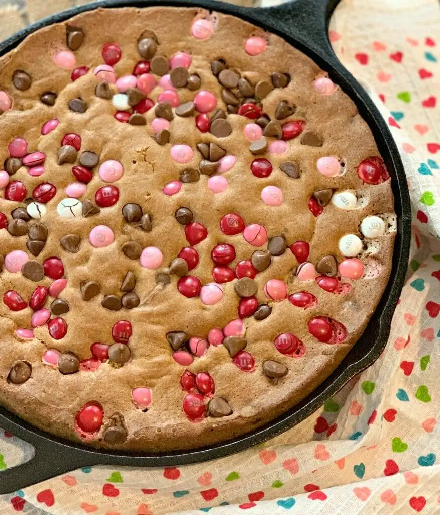 A festive cookie skillet loaded with pink, red, and white candy-coated chocolates and chocolate chips, baked to golden perfection, perfect for sharing on a cozy Valentine's Day.