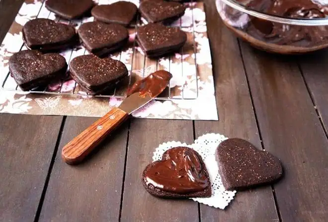 Rich chocolate heart cookies cooling on a wire rack, with one cookie in the foreground being generously spread with creamy chocolate frosting, perfect for a decadent Valentine's Day dessert.