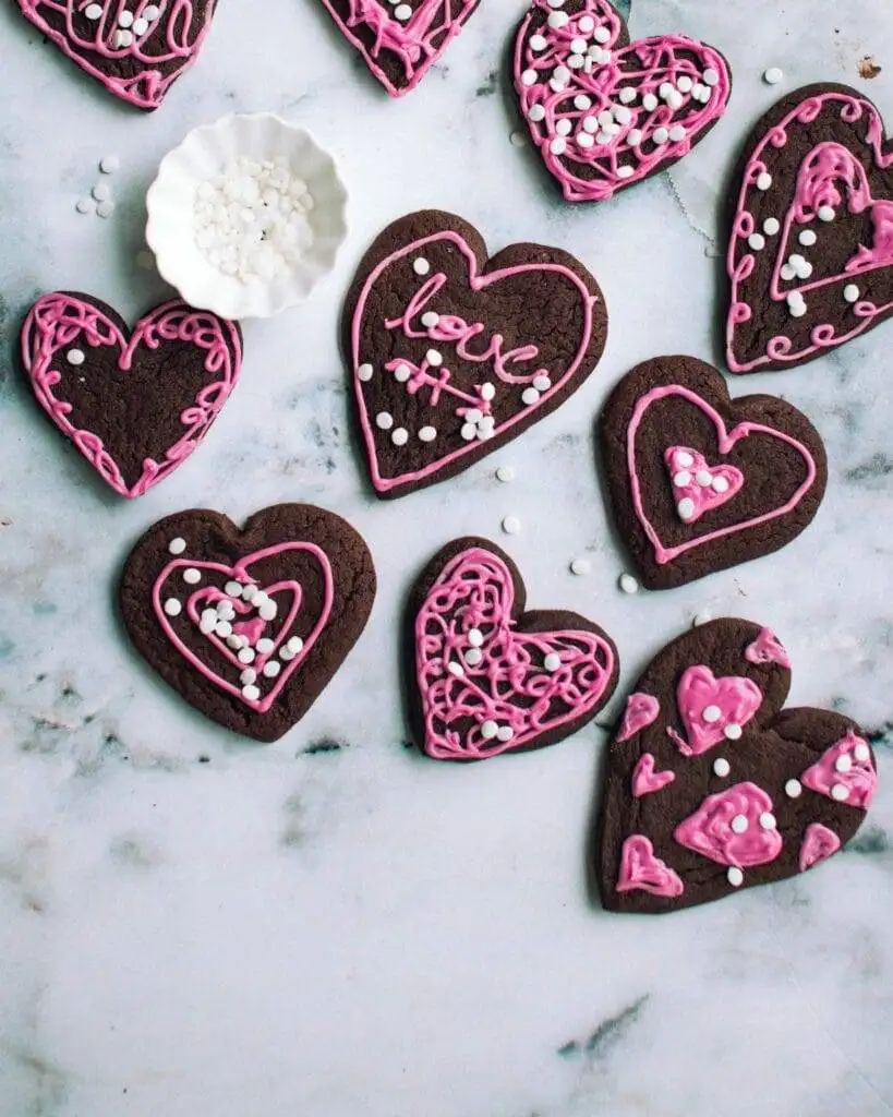 Chocolate heart cookies decorated with pink icing patterns and white sprinkles, arranged on a marble surface alongside a small bowl of extra sprinkles, perfect for a festive and romantic treat.
