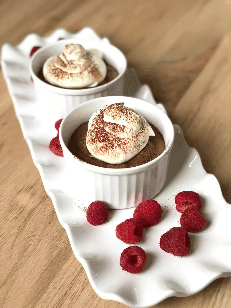 Two ramekins of rich chocolate mousse topped with whipped cream and dusted with cocoa, served on a white scalloped tray with fresh raspberries, perfect as romantic desserts for two.