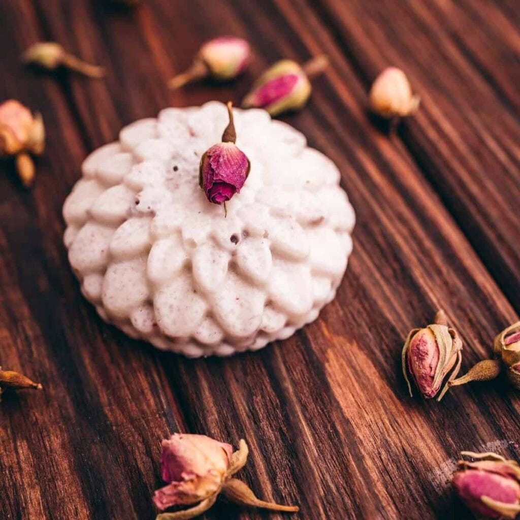 A beautifully crafted white DIY bath bomb with a floral texture, topped with a dried rosebud and surrounded by scattered rosebuds on a rustic wooden surface. A charming idea for DIY Valentine's Crafts, offering a thoughtful and handmade gift option.