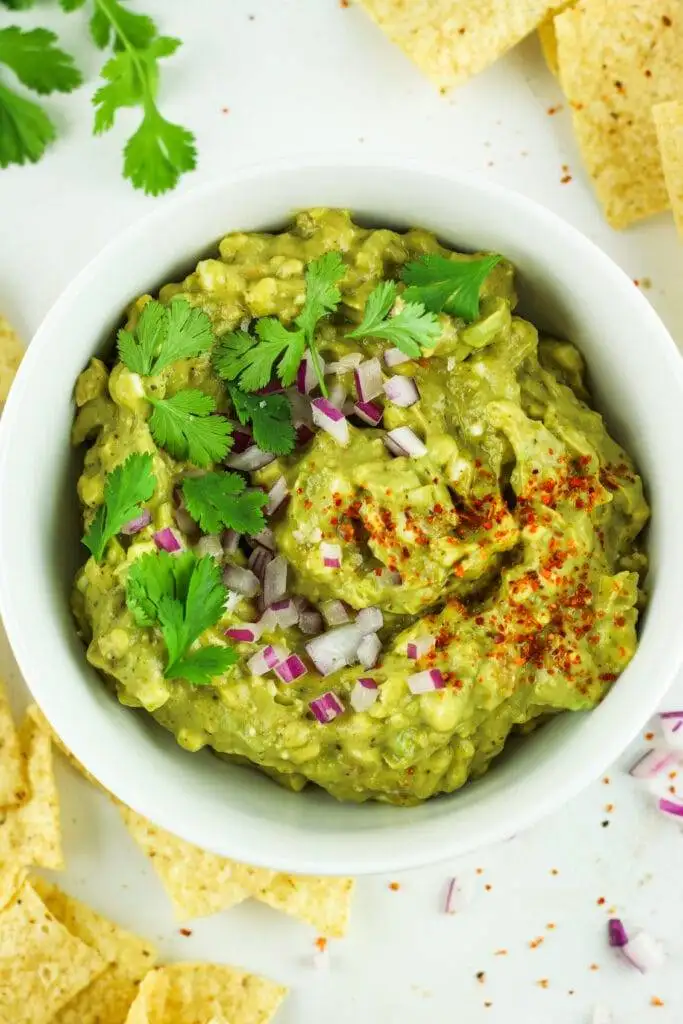 A vibrant bowl of creamy guacamole topped with fresh cilantro leaves, diced red onions, and a sprinkle of chili powder, served with tortilla chips. Perfect for enjoying as one of the best party dips for any occasion.