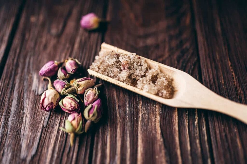 A wooden scoop filled with homemade sugar scrub, featuring visible grains and small dried rose petals, placed on a rustic wooden surface alongside a cluster of dried rosebuds. A thoughtful and elegant idea for DIY Valentine's Crafts, perfect for creating personalized self-care gifts.