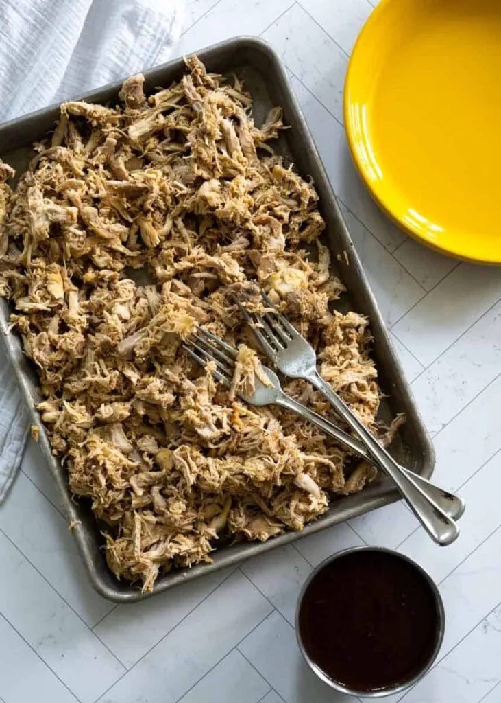 A tray of shredded pulled pork with two forks, served alongside a bowl of barbecue sauce. A versatile and savory Super Bowl food idea perfect for sandwiches or sliders.