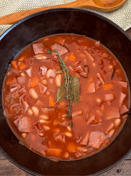 Rich and savory ham and bean stew in a black cast-iron pot, featuring hearty chunks of ham, white beans, carrots, and a garnish of fresh thyme and bay leaf.