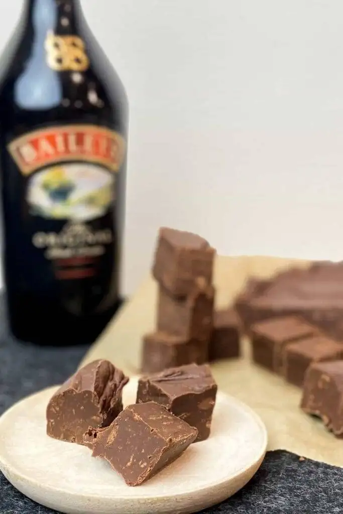 A close-up of rich and creamy fudge made with Baileys Irish Cream, cut into bite-sized pieces and displayed on a wooden plate. A bottle of Baileys sits in the background, emphasizing the indulgent, Irish-inspired treat. Perfect for St. Patrick's Day Food Ideas and adding a sweet touch to Irish Recipes.