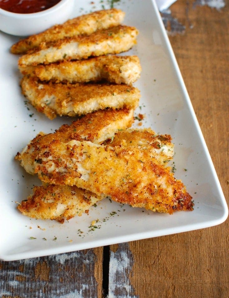 A plate of crispy baked chicken tenders coated in a golden breadcrumb crust, garnished with parsley and served with a side of dipping sauce. These flavorful and kid-friendly treats are a classic option for Superbowl appetizers.
