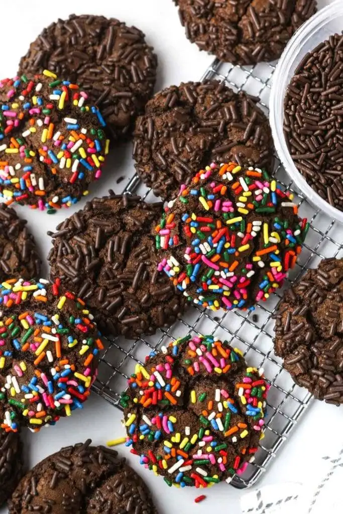 Chewy chocolate heart cookies covered in colorful rainbow sprinkles and chocolate jimmies, arranged on a cooling rack for a festive and fun treat.