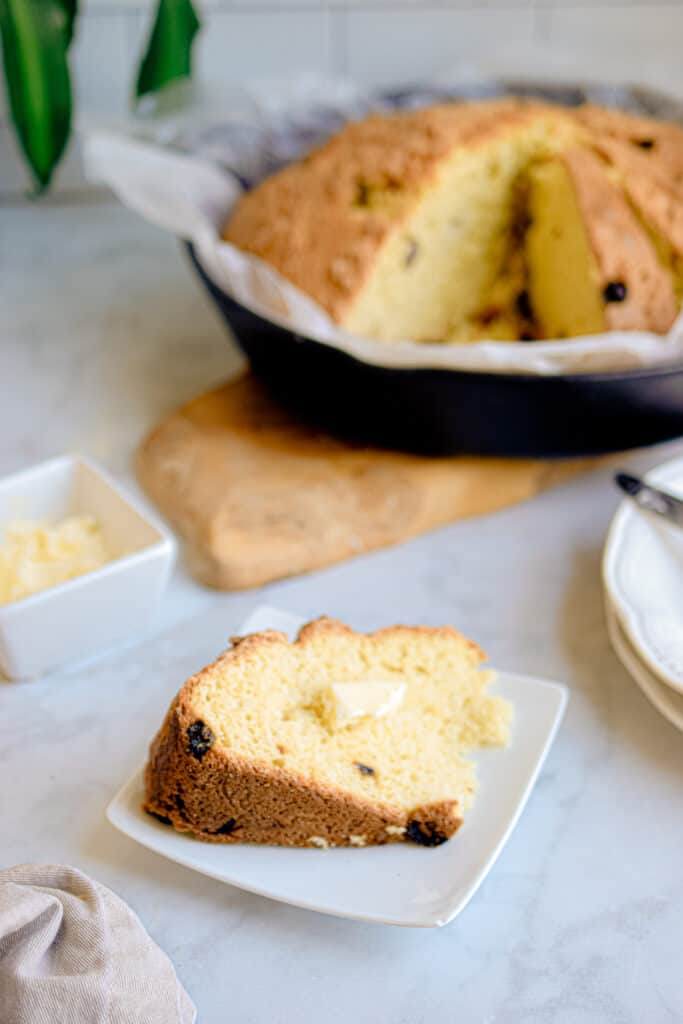 Slice of Irish soda bread with raisins served on a white plate, topped with a pat of butter, with the remaining loaf in the background—perfect for St. Patrick's Day food ideas and traditional Irish recipes.