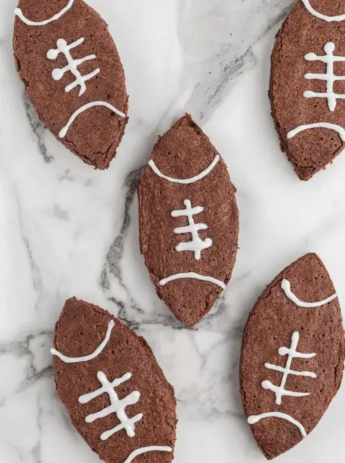 Football-shaped chocolate brownies decorated with white icing laces, arranged on a marble surface—fun and festive treats perfect for a game day or Super Bowl party.