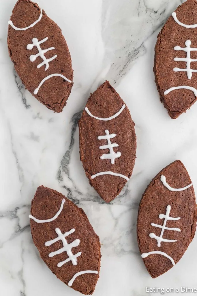 Football-shaped brownies decorated with white icing laces, arranged on a marble surface. A festive and chocolatey Super Bowl food idea for game day treats.