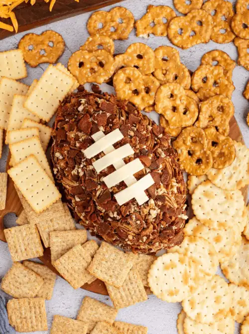 Football-shaped cheese ball coated in bacon and pecans, topped with cheese laces, and surrounded by assorted crackers—perfect centerpiece for Super Bowl food ideas.