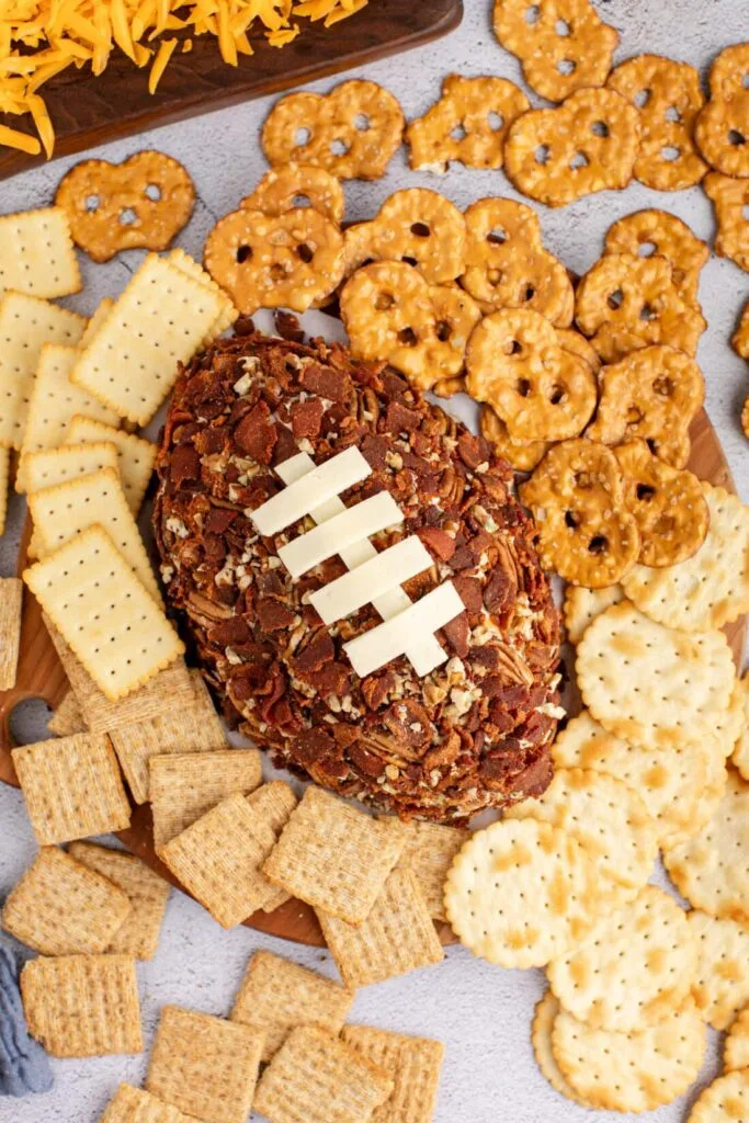 Football-shaped cheese ball coated with chopped pecans, topped with cheese laces, surrounded by an assortment of crackers and pretzels. A festive and crowd-pleasing Super Bowl food idea for game day gatherings.