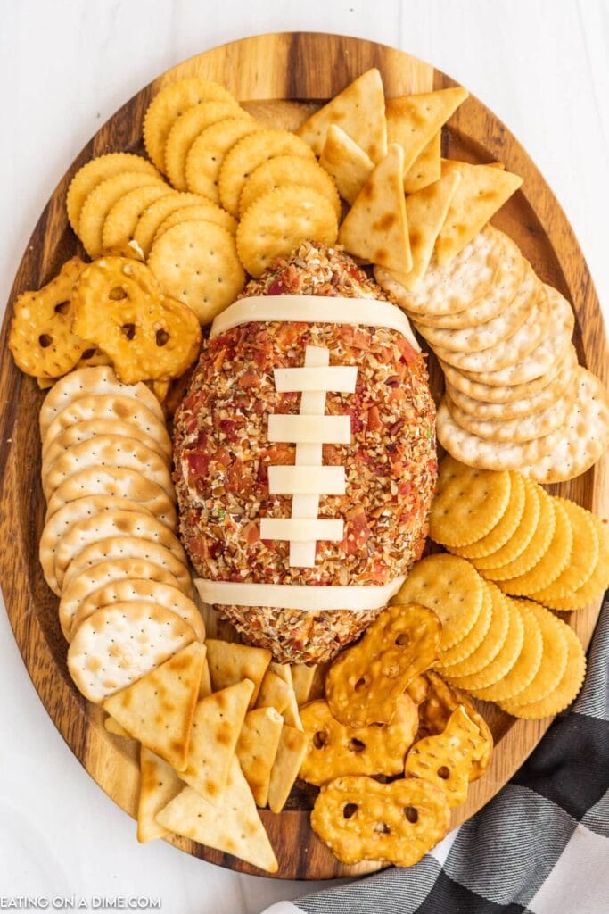 A creative Superbowl appetizer featuring a football-shaped cheese ball topped with chopped nuts and bacon, decorated with strips of cheese to resemble football laces. Surrounded by an assortment of crackers and pretzels, presented on a wooden serving tray, perfect for game day celebrations.