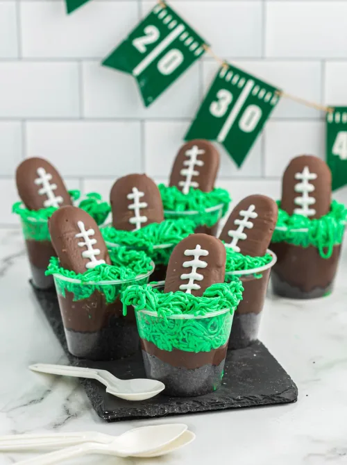 Individual chocolate pudding cups topped with green frosting “grass” and football-shaped cookies decorated with white icing laces, perfect for a football-themed party or Super Bowl dessert table.