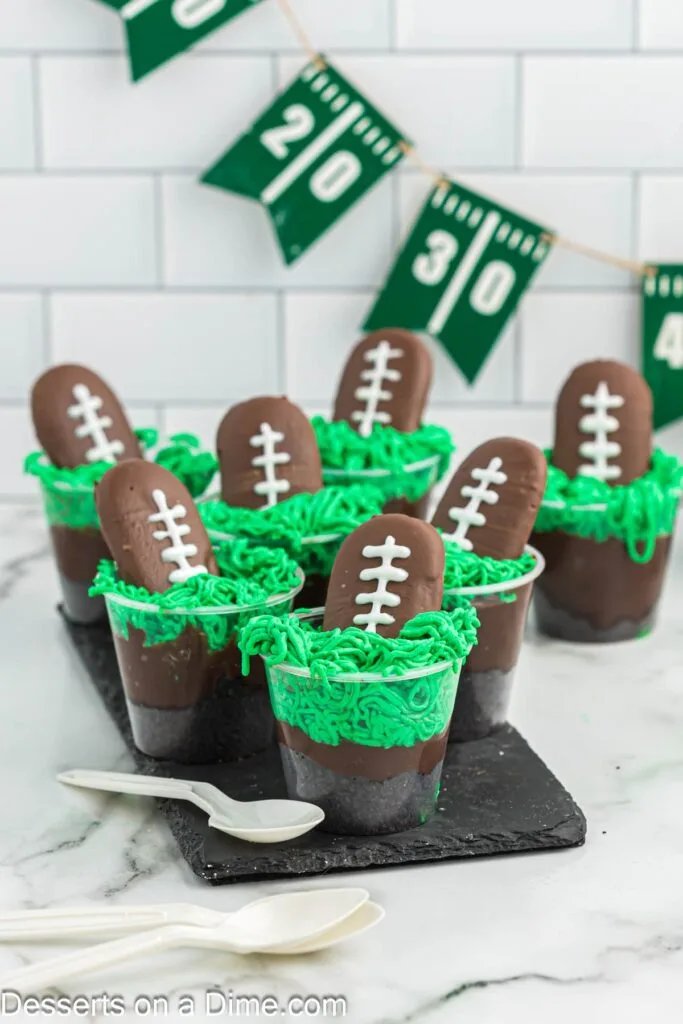 Football-themed pudding cups topped with green frosting to resemble grass and decorated with football-shaped cookies. A creative and fun Super Bowl food idea for game day desserts.