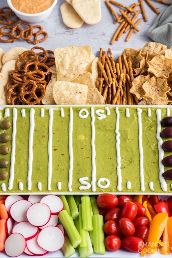 A fun Superbowl appetizer platter designed like a football field, featuring guacamole spread as the field with sour cream piped to create yard lines. The platter is surrounded by chips, pretzels, and crackers, along with fresh veggies like celery, radishes, cherry tomatoes, and olives, providing a mix of healthy and indulgent snack options.