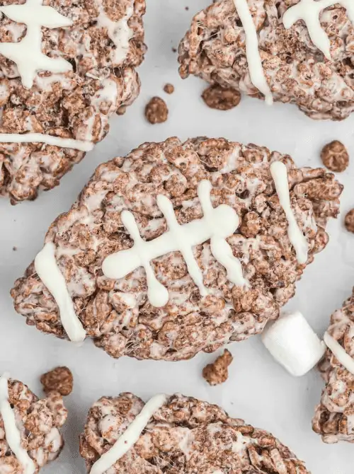 Chocolate crispy rice treats shaped like footballs, decorated with white icing laces and marshmallow ends—fun and festive desserts for game day celebrations.