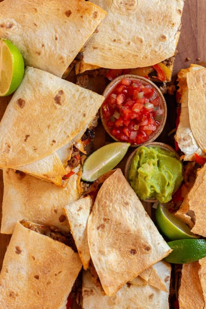 A platter of golden-brown quesadillas filled with seasoned meat, cheese, and sautéed vegetables, served with sides of fresh guacamole, salsa, and lime wedges. Perfect for sharing, this dish is a delicious Superbowl appetizer for game day gatherings.