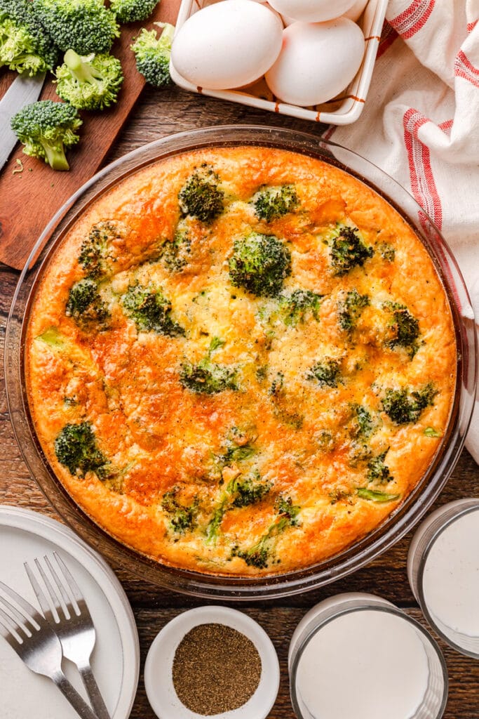 Freshly baked broccoli and cheese quiche, perfect for quick meals with ham, surrounded by ingredients like fresh broccoli, eggs, and seasoning on a rustic wooden table.