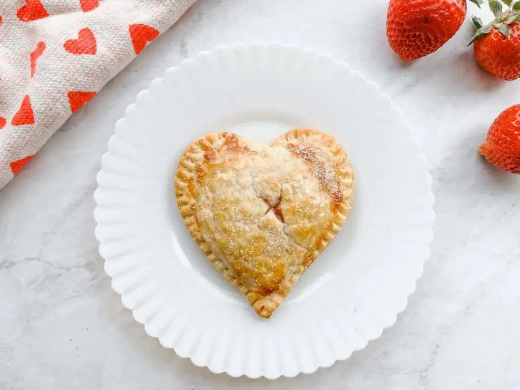 A heart-shaped hand pie dusted with sugar, baked to golden perfection and served on a white plate, ideal for romantic desserts for two or a charming Valentine’s Day treat.