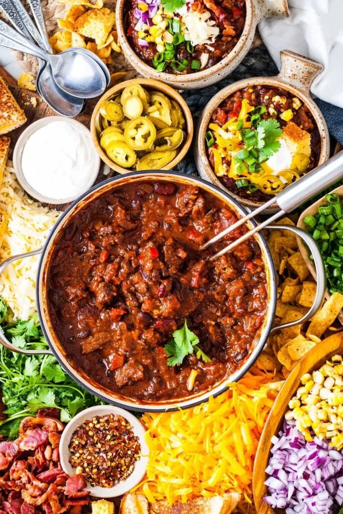 A vibrant chili bar setup featuring a pot of hearty chili surrounded by toppings like shredded cheese, jalapeños, sour cream, diced onions, and fresh cilantro. A customizable and crowd-pleasing Super Bowl food idea for game day gatherings.