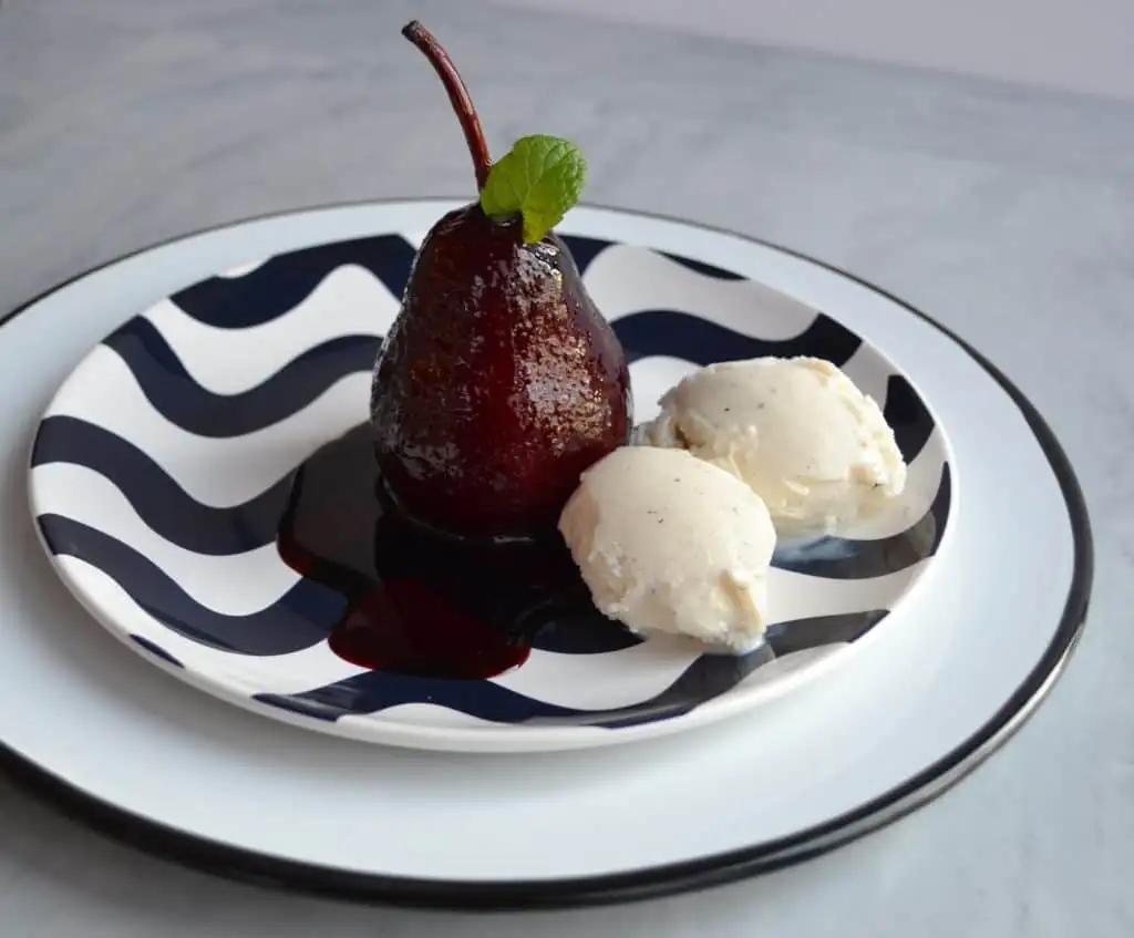A beautifully plated poached pear in red wine sauce, garnished with a fresh mint leaf, served alongside two scoops of vanilla ice cream, making it an elegant and indulgent option for romantic desserts for two.