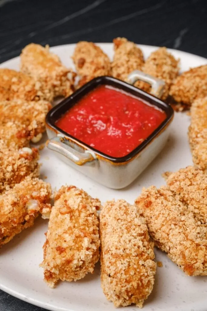 Crispy baked mozzarella sticks arranged in a circle on a white plate, with a square dish of marinara sauce in the center. A classic and budget-friendly Super Bowl food idea for game day snacking.