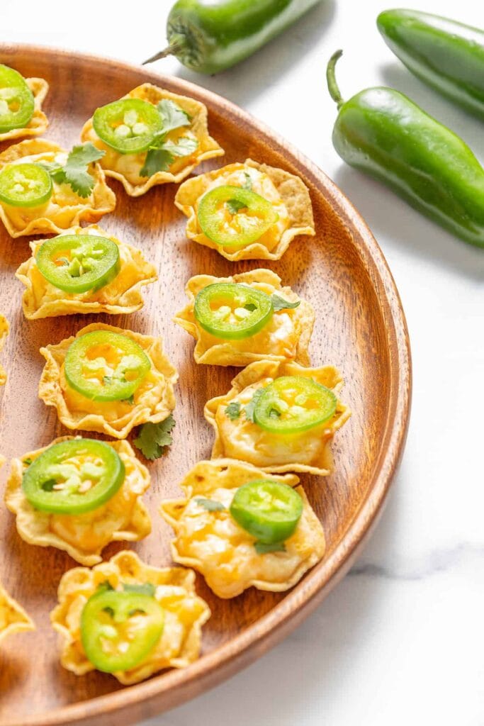 A wooden platter filled with bite-sized nacho cups made from tortilla chips, topped with melted cheese and sliced jalapeños, garnished with fresh cilantro. Perfect for spicy and crunchy Superbowl appetizers.