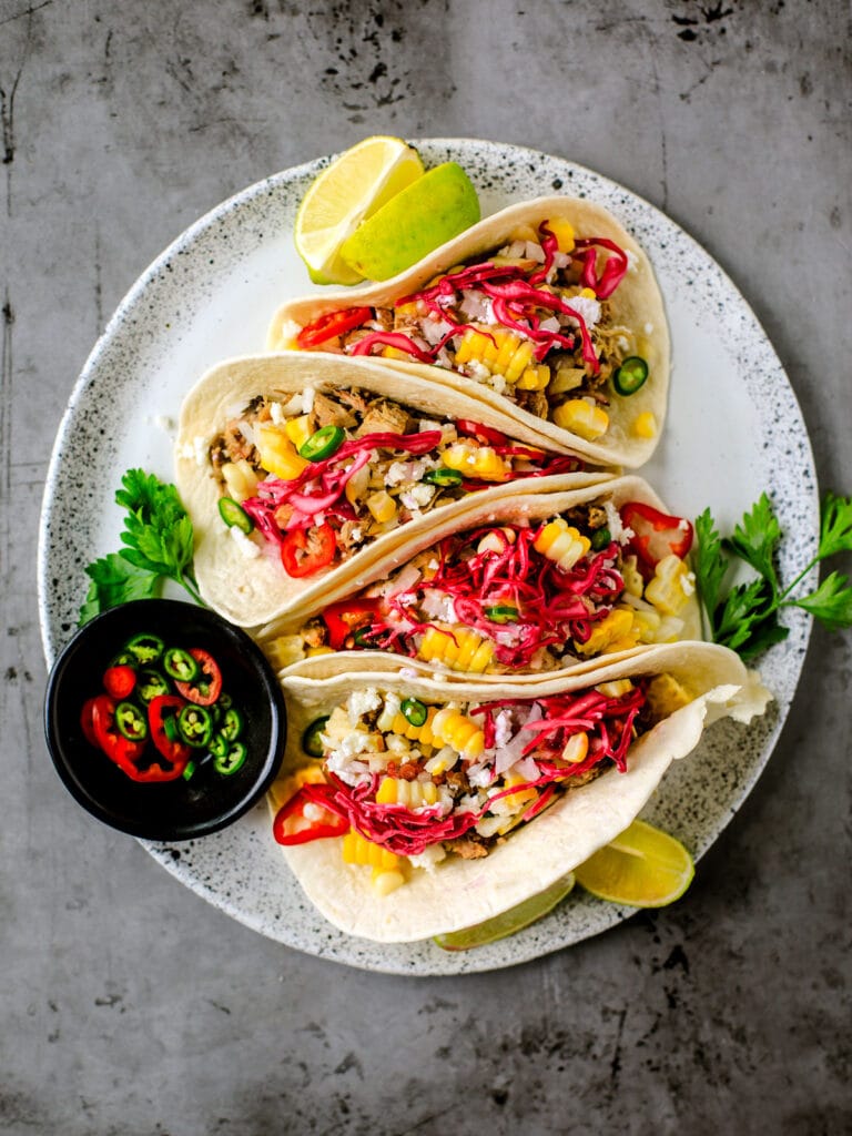 Fresh tacos filled with leftover pulled pork, topped with pineapple salsa, pickled red cabbage, sliced chili peppers, and garnished with lime wedges on a speckled plate.