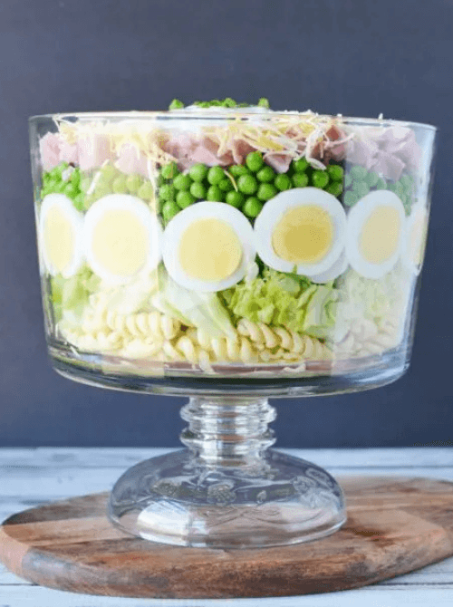 Layered pasta salad in a glass trifle bowl, featuring quick meals with ham, hard-boiled eggs, peas, lettuce, and shredded cheese, displayed on a wooden board.