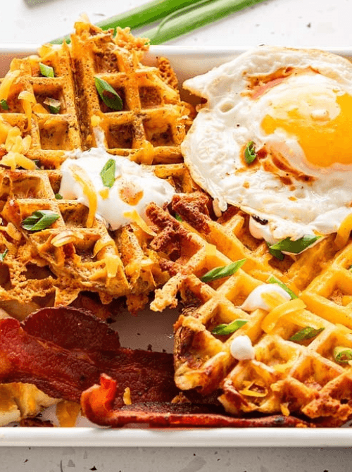 Plate of crispy savory waffles made from leftover ingredients, topped with a fried egg, shredded cheese, sour cream, and green onions—an inventive and delicious twist for creative leftovers recipes.